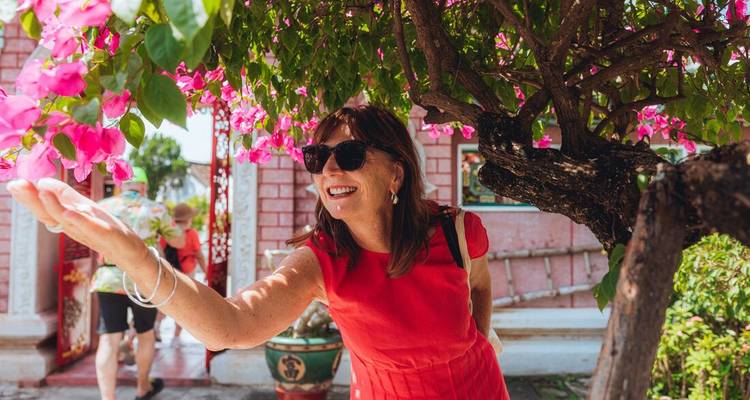 A woman in a red dress reaches out to touch bright pink bougainvillea blossoms beside historic architecture.