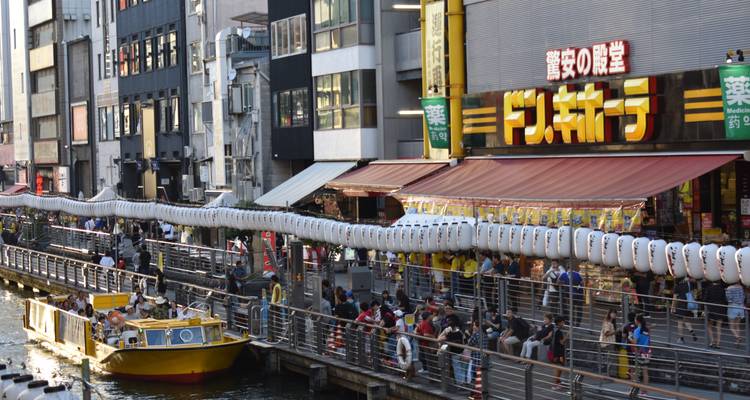 Canal animé de Dotonbori à Osaka bordé de lanternes, de restaurants et d'un bateau touristique jaune qui passe.