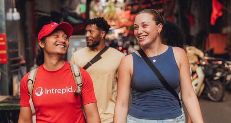 Trois voyageurs se promènent dans un marché de rue animé du Vietnam, souriant et discutant avec des scooters et des étals en arrière-plan.