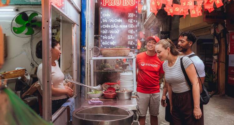 Les voyageurs se pressent autour d'un étal de pho fumant dans une ruelle étroite, discutant avec le vendeur local sous un panneau de menu en néon.
