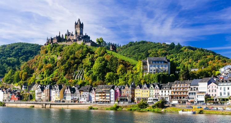 Vue panoramique du château de Reichsburg Cochem perché sur une colline verdoyante au-dessus de maisons riveraines colorées le long de la Moselle sous un ciel bleu éclatant.