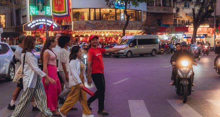 Scène de rue en soirée alors qu'un guide vêtu d'une chemise rouge guide un groupe à travers un carrefour très fréquenté illuminé par des enseignes lumineuses.