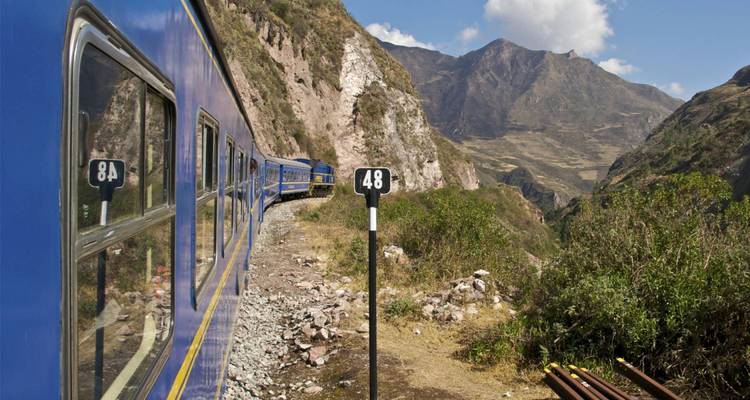 Blue PeruRail trein die langs een bergkloof kronkelt met ruige Andespico's boven je hoofd.