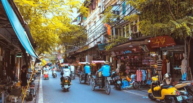 Marché de rue animé avec des gens sur des scooters et des pousse-pousse sous des arbres feuillus.