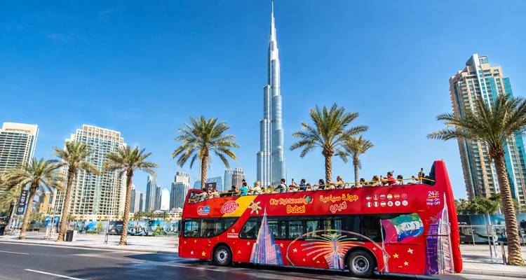 Een felrood open-top toerbus rijdt langs palmbomen met de Burj Khalifa die hoog uittorent tegen een hemelsblauw.
