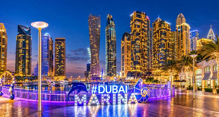Vista nocturna del horizonte de Dubai Marina iluminado en púrpura vívido y oro con un letrero brillante de #Dubai Marina en primer plano.