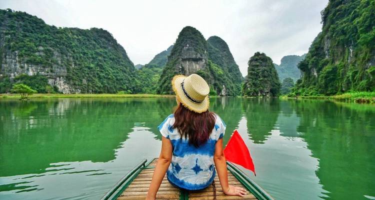 Rückansicht einer Frau auf einem hölzernen Sampan, das durch smaragdgrünes Wasser zwischen hoch aufragenden Karstgipfeln gleitet