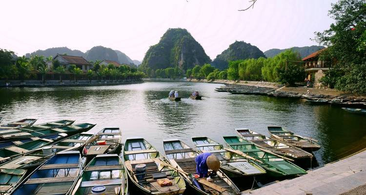 Reihe traditioneller Ruderboote am Fluss vertäut mit Karstbergen und palmengesäumtem Dorf im Hintergrund