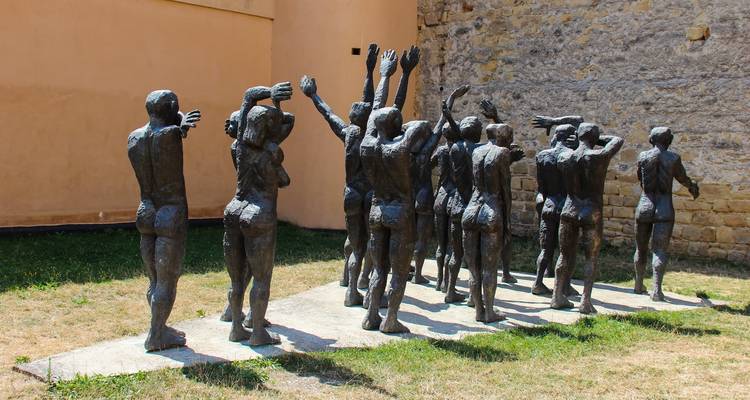 Sculpture de bronze sombre représentant des figures émaciées les bras levés contre un mur nu
