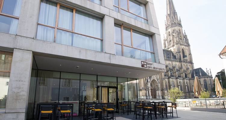 Extérieur d'un hôtel moderne en verre situé à côté de la Cathédrale de Linz avec des places assises extérieures sur la terrasse.