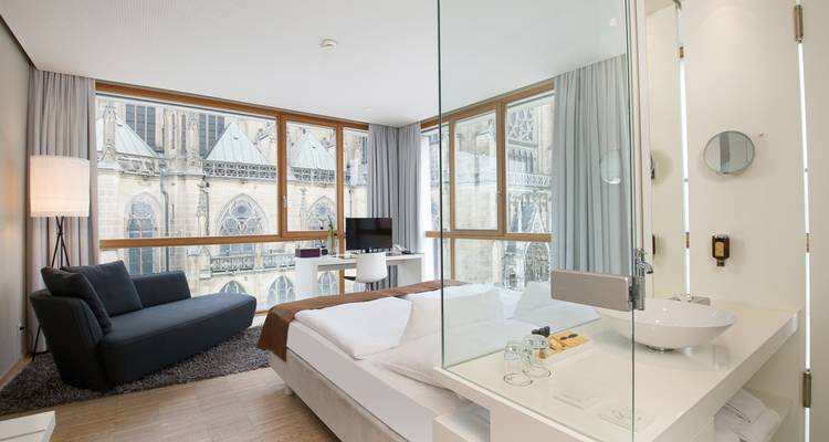 Chambre d'hôtel minimaliste avec salle de bains aux parois vitrées, grandes fenêtres encadrant l'architecture de la cathédrale à l'extérieur.