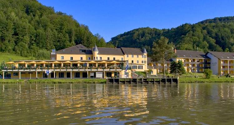 Hôtel resort en bord de rivière avec façade jaune et terrasses reflétées dans une eau calme sur fond de forêt.