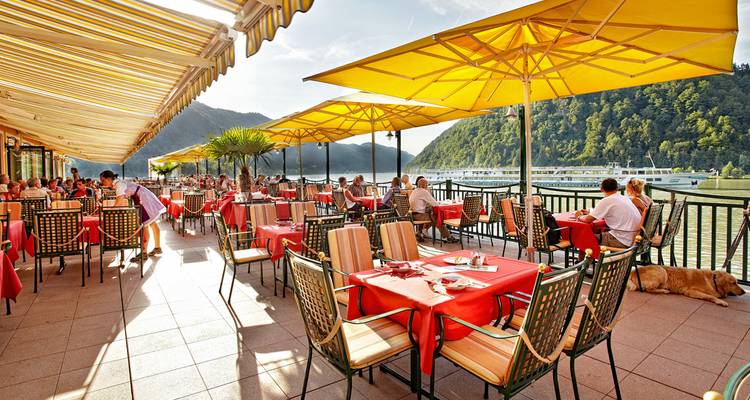 Restaurant de terrasse ensoleillée avec des nappes rouges donnant sur un méandre de rivière et des collines boisées, clients savourant leurs repas.