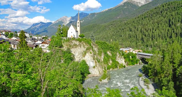 L'église blanche sur la colline surplombe une rivière alpine rapide et un pont en bois couvert encadré par les pics.