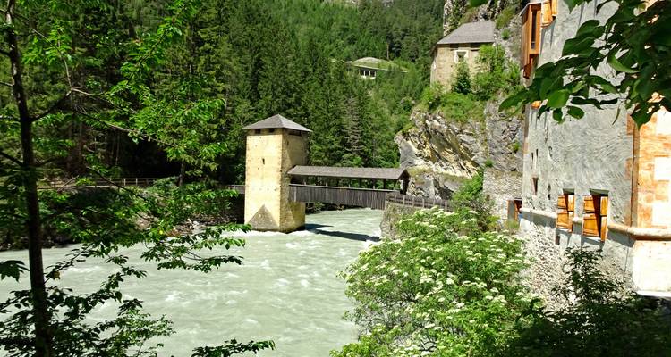 Une tour en pierre historique et un pont couvert enjambent une rivière alimentée par un glacier dans une gorge forestière.