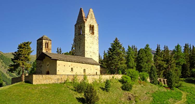 Église en pierre historique avec deux tours inégales perchées sur une colline herbeuse entourée de sapins, se dressant sur un ciel bleu dégagé.