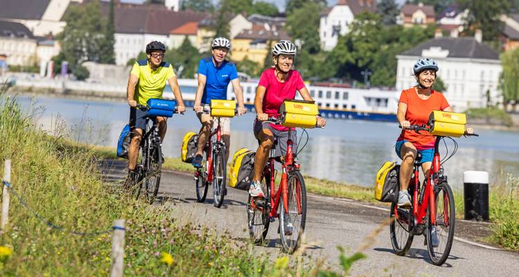 Ligne de cyclistes circulant le long d'une piste riverside avec des bâtiments de la ville et des bateaux amarrés à l'arrière.