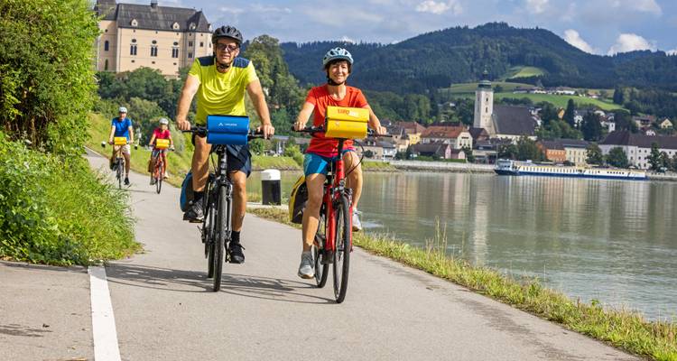Deux cyclistes de face qui passent devant une ville riveraine avec un château historique et une église sur la colline.