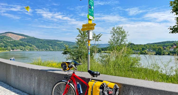 Vélo de randonnée rouge avec sacoches jaunes appuyé contre un mur bas dominant les panneaux de signalisation de la piste cyclable du Danube.