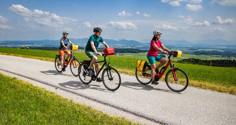 Three cyclists follow a narrow country road through rolling green farmland under a blue sky.