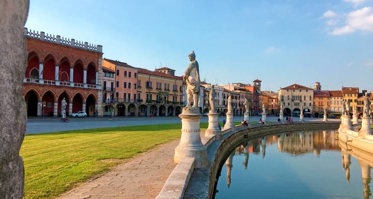 Les élégantes statues de la Piazza Prato della Valle et ses loggias en arc se reflètent dans le canal tranquille sous la lumière chaude du soir à Padoue.