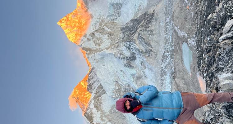 Bundled trekker at dawn stands on a rocky ridge with sun-kissed Everest peaks blazing orange behind.