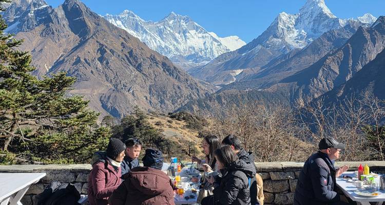 Travellers share outdoor breakfast on a stone terrace with panoramic snowy Himalayan peaks towering beyond.