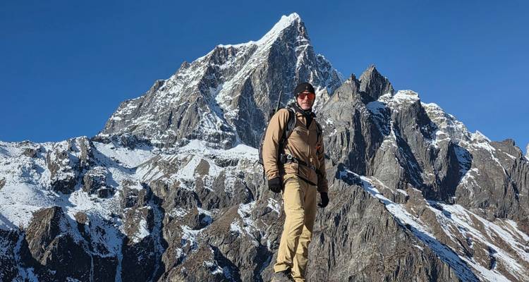 Solo hiker poses against a sharp snow-covered Himalayan spire under a deep blue sky.