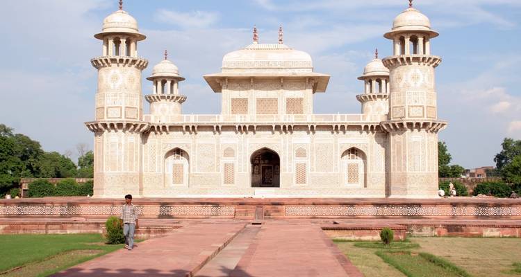 Das kunstvolle weiße Marmormausoleum Itimad-ud-Daulah, bekannt als das Baby Taj, steht symmetrisch vor einem blauen Himmel, während ein einzelner Besucher in der Nähe spaziert.