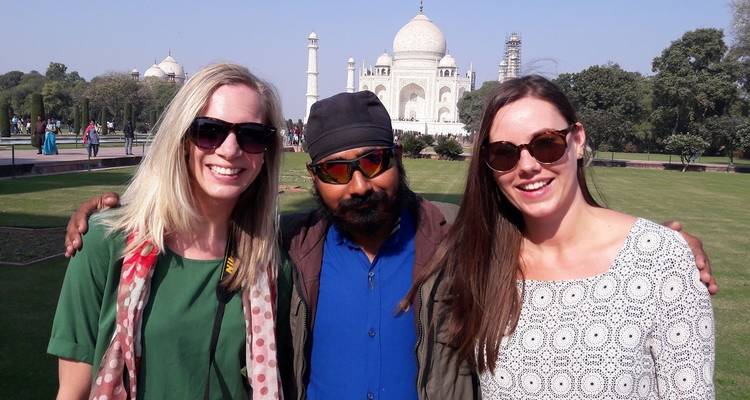 Zwei Frauen und ein lokaler Führer stehen Arm in Arm lächelnd vor dem sonnenbeschienenen Taj Mahal und seinen Gärten.