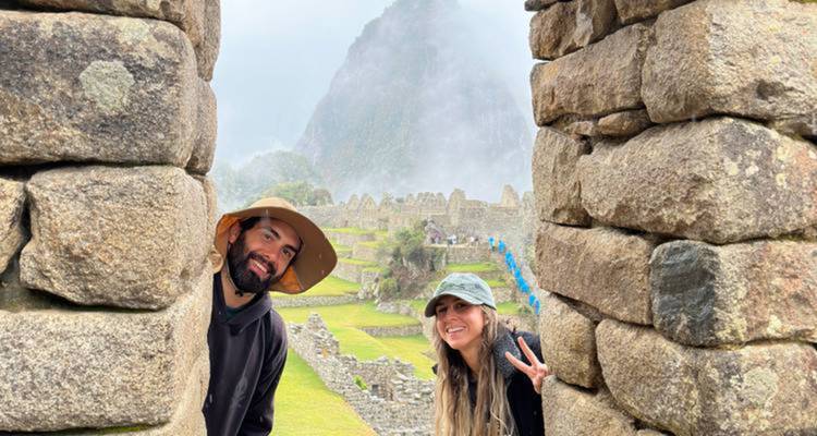 Dos personas asomándose desde detrás de ruinas incas con montañas brumosas.
