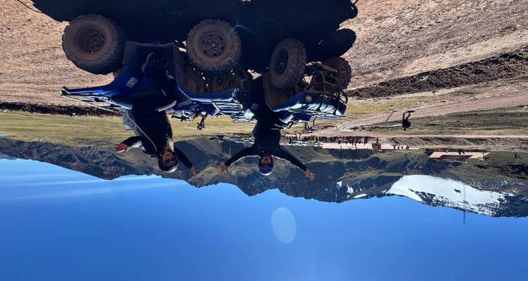 Deux voyageurs sur des quads bleus lèvent les bras sur un sentier de terre avec en arrière-plan des montagnes andines aux pics enneigés sous un ciel bleu éclatant.