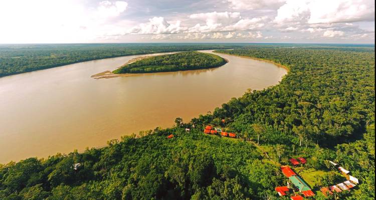 Panorama aéreo del ancho río Amazonas de color marrón curvándose a través de la infinita selva tropical verde con un albergue de techo rojo abajo