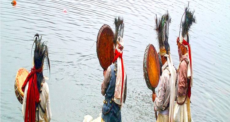 Vier Menschen in traditioneller Kleidung halten Trommeln in der Nähe eines Gewässers.