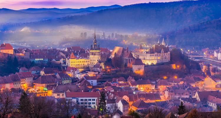 Evening panorama of Sighisoara’s historic citadel glowing with lights amid rolling hills and soft mist.