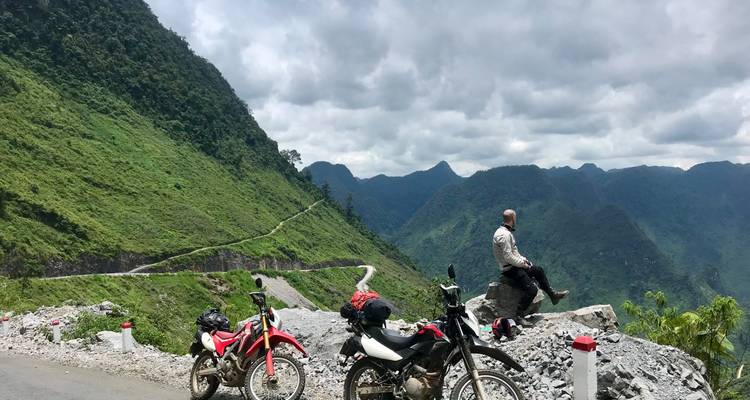 Hombre sentado en una roca con motocicletas y paisaje montañoso a la vista.