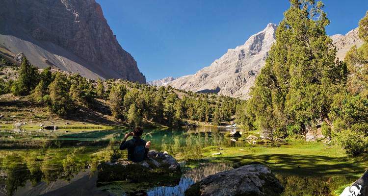 Bergige Seenlandschaft mit einer Person, die auf einem Felsen sitzt.