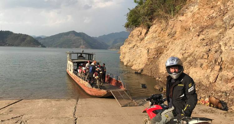Motorrijder op een steiger met een boot en bergen op de achtergrond.