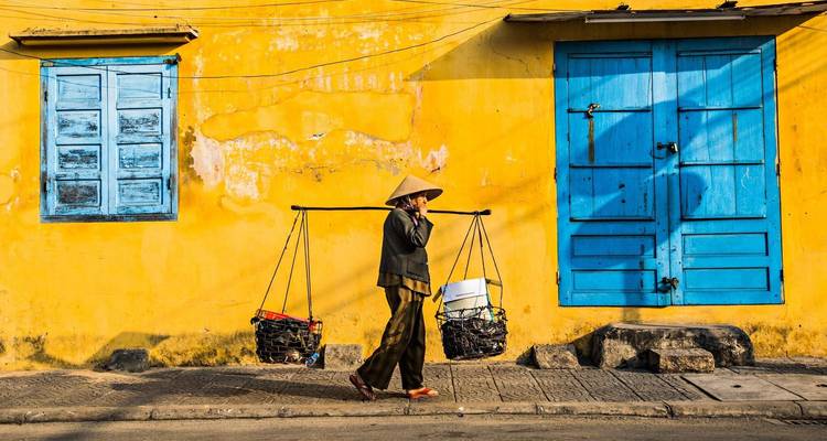 Person, die Körbe an einer Stange vor einer gelben Wand mit blauen Fensterläden trägt.