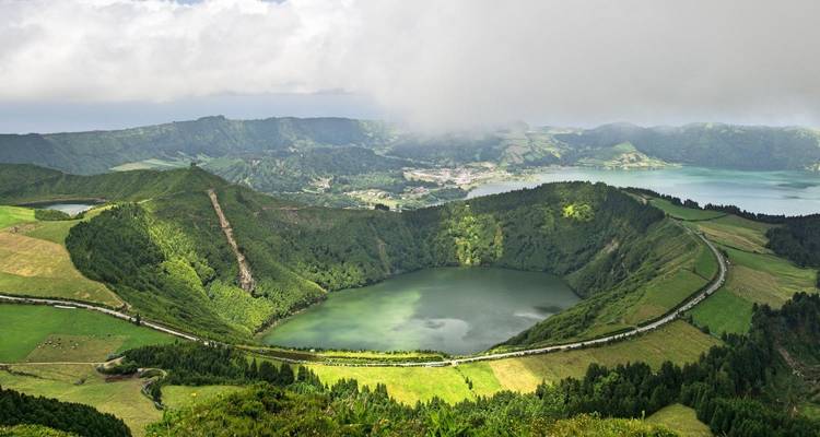 Vue aérienne de Sete Cidades aux cratères jumeaux avec ses lacs émeraude entourés de crêtes volcaniques luxuriantes et de terres agricoles en damier sous un ciel brumeux.