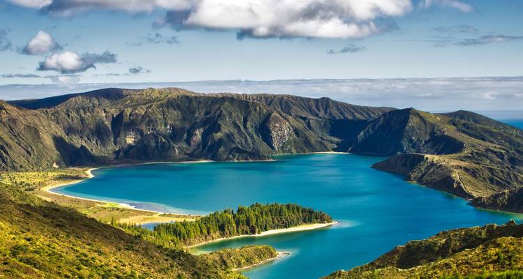 Les eaux turquoise de la Lagoa do Fogo entourées de montagnes volcaniques vertes et escarpées et d'une petite péninsule boisée sous un ciel partiellement nuageux.