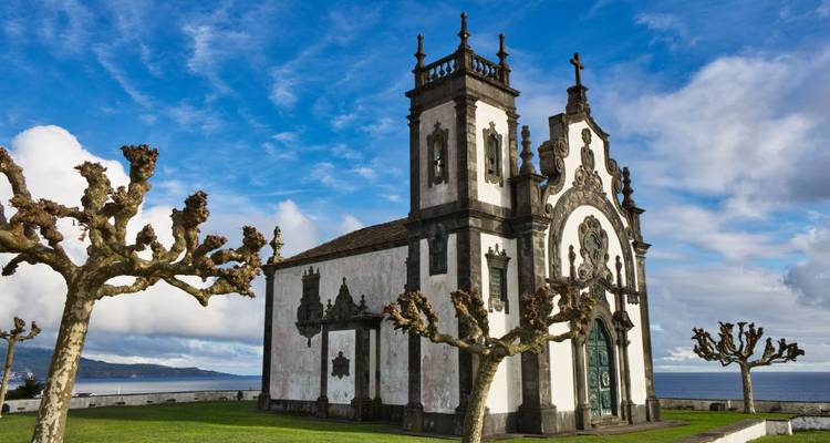 Chapelle côtière historique aux couleurs blanc et noir, dotée d'une façade ornée, se détachant sur un ciel bleu et l'océan, encadrée par des arbres taillés sur une terrasse herbeuse.