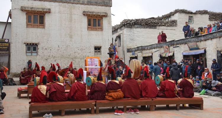 Monks in red robes sit in a courtyard watching a traditional festival performance in a Himalayan village.