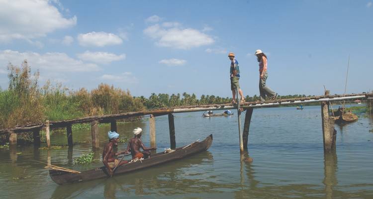 Menschen überqueren eine Holzbrücke über einen Fluss, während Kanus darunter vorbeifahren.