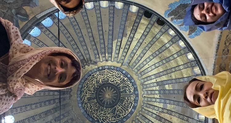 Vue en contre-plongée de quatre visiteurs souriants autour du bord d'un grand dôme ottoman décoré de carreaux bleus et de calligraphie arabe.