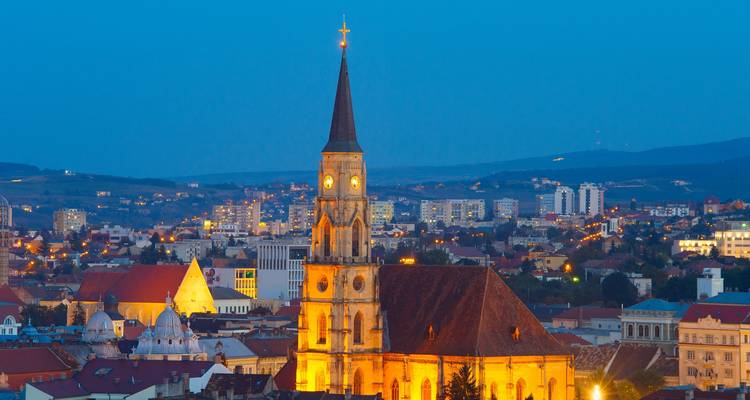 Blue-hour panorama of Cluj-Napoca with illuminated St. Michael's Church spire above the city lights.