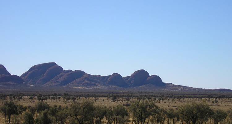 Distant silhouette of Kata Tjuta domes seen across flat arid plain under clear blue sky.
