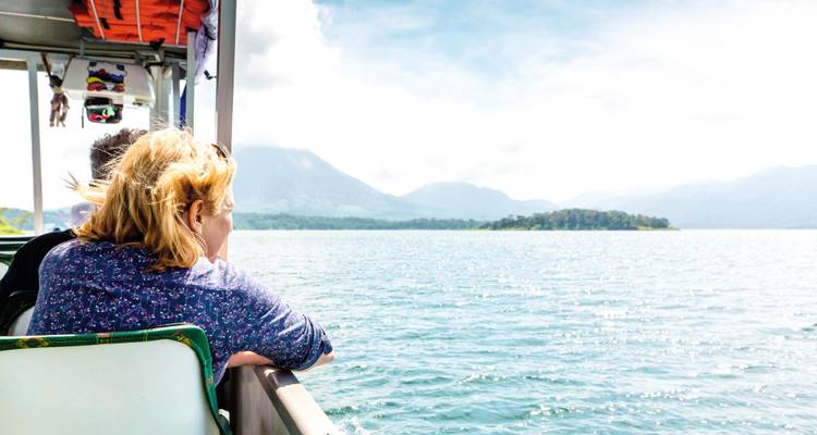 Un passager s'appuie sur la rambarde d'un bateau de lac, admirant des volcans brumeux et des rives couvertes de forêt tropicale.