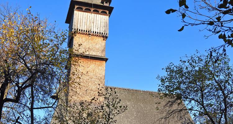 La haute tour d'église en bois s'élève au-dessus des arbres d'automne sous un ciel bleu profond.