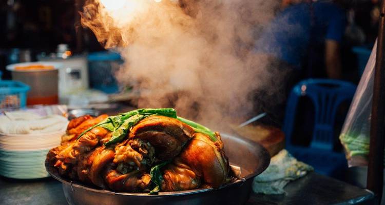 Un wok fumant rempli de jarrets de porc braisés et de légumes verts dans un étal de restauration nocturne animé.